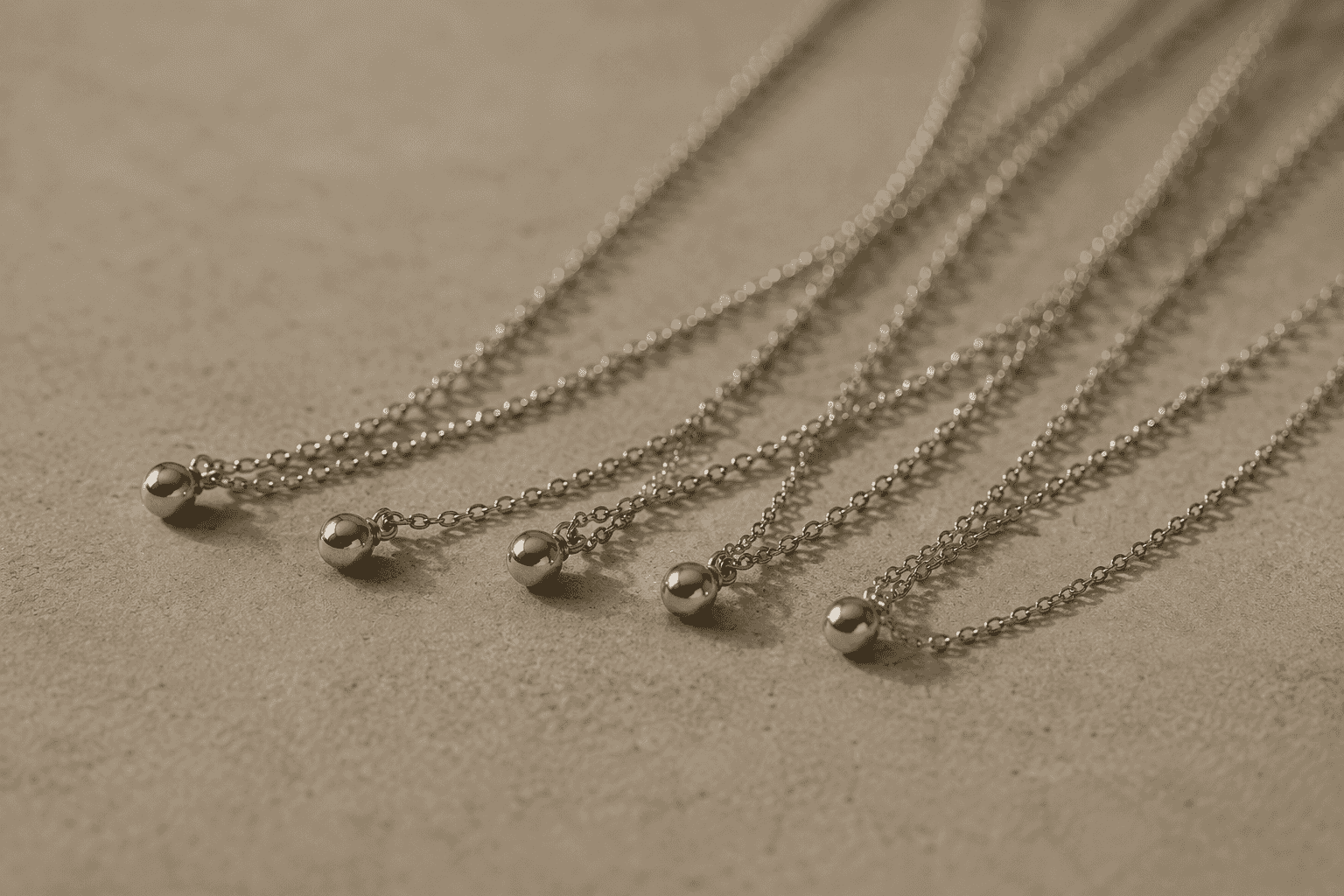 Several fine silver chains, each ending in a small ball pendant, laid loosely across pale stone.