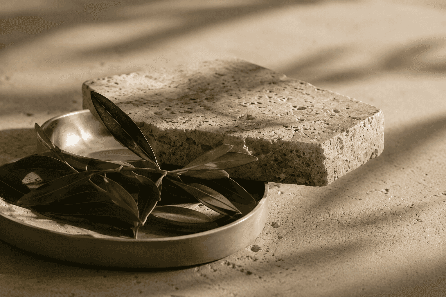 A textured travertine stone slab resting on a metal dish, with an olive branch tucked beneath it.