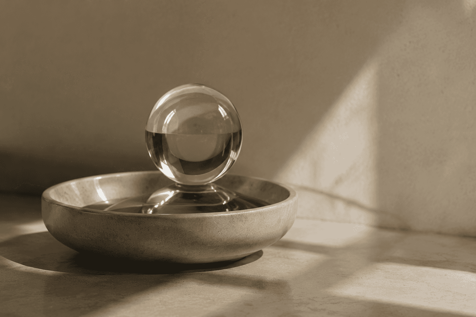 A clear glass sphere balanced atop a shallow ceramic dish, photographed against a plain warm background.