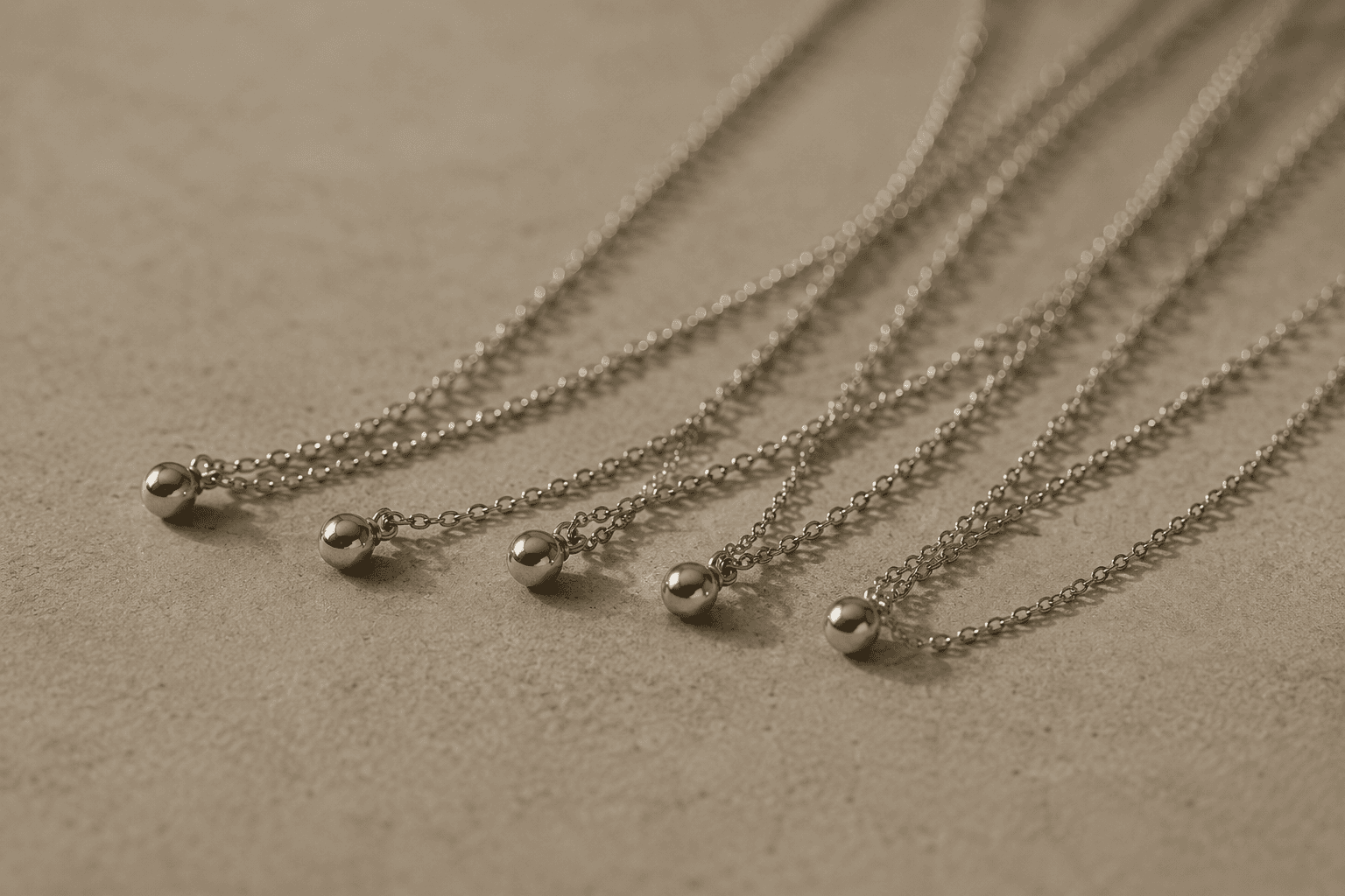 Several fine silver chains, each ending in a small ball pendant, laid loosely across pale stone.
