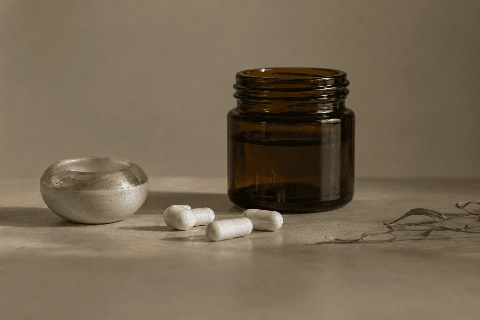 White capsules beside a small marble bowl and an amber glass jar, with olive leaves at the edge.