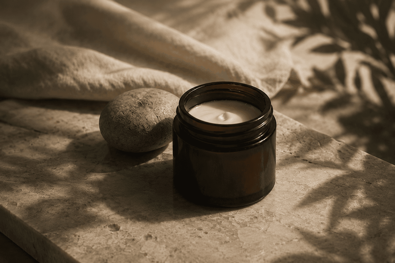 An open jar of cream beside a smooth stone and folded linen, photographed in warm afternoon light.