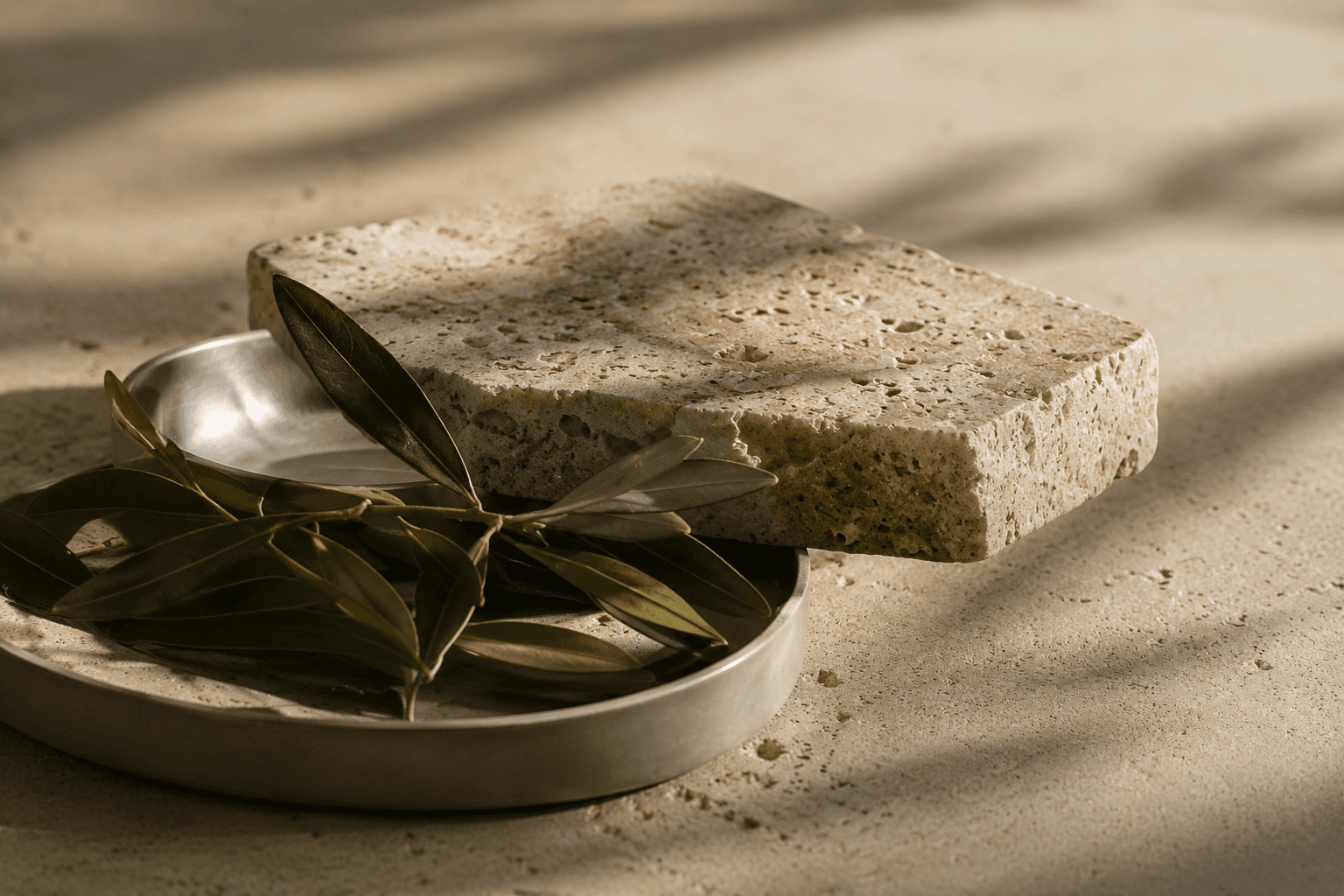 A textured travertine stone slab resting on a metal dish, with an olive branch tucked beneath it.